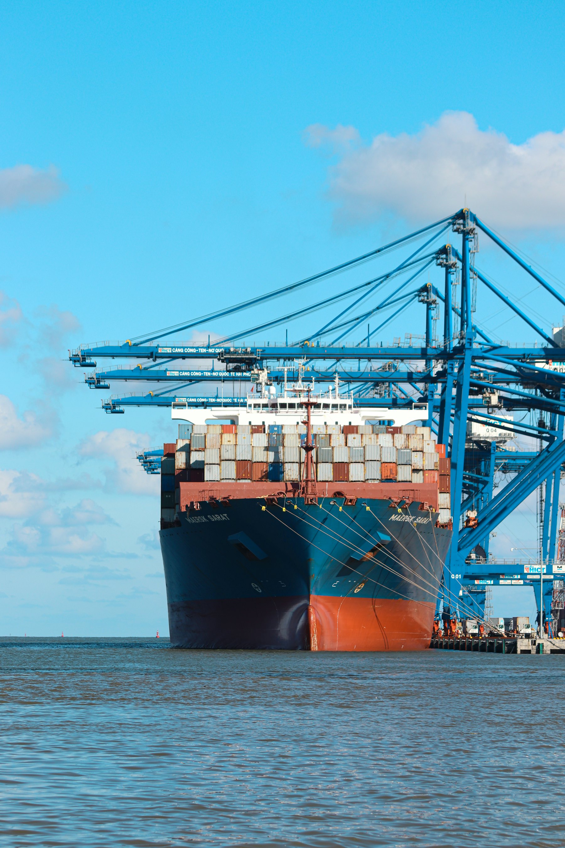 Container ship docked beside blue port cranes under a clear sky
